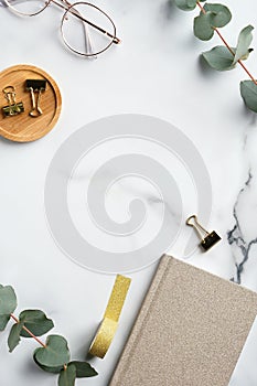 Modern marble office desk table with paper notebook  glasses  supplies and eucalyptus leaves.