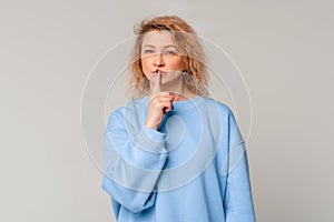 Modern mature woman with curly blonde hair showing shhh secret hush gesture, shushing, stands in trendy blue sweatshirt over light