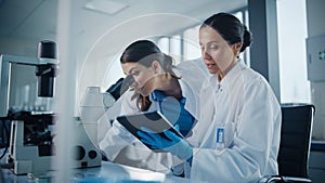 Modern Medical Research Laboratory: Two Female Scientists Working Together Using Microscope, Analy
