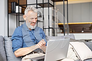 Modern middle-aged bearded man using laptop computer sitting on the couch at home