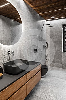Modern minimalist bathroom interior with stone grey tiles, wooden furniture, black sink and toilet and round mirror.