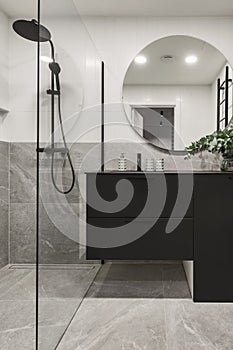 Modern minimalistic bathroom interior design with grey stone tiles, black furniture, eucalyptus in glass vase, round mirror.