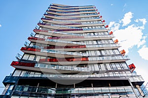 A modern multi-story building with staggered balconies featuring vibrant red accents, set against a clear sky with wispy