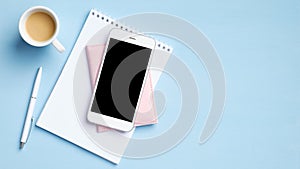 Modern office desk with smartphone with blank mockup screen, cup of coffee, blank paper notepad, document on blue background. Top