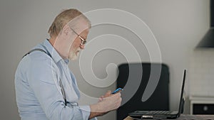 Modern old age, busy elderly man in glasses paying bills online uses laptop and a mobile phone while sitting at desk in