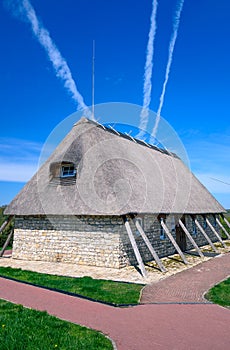 A modern old-looking house with a thatched roof and stone walls and support beams on its sides. Paved path in front and blue sky