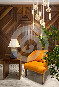 Modern orange armchair, lamp shade on small wooden table, contemporary tall glass chandelier, and wood cladding wall.