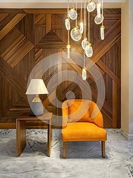 Modern orange armchair, lamp shade on small wooden table, contemporary tall glass chandelier, and wood cladding wall.