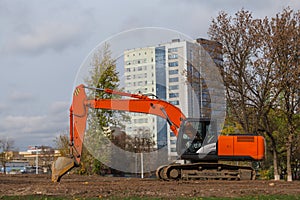Modern orange excavator with a lowered bucket stands on a cleared construction site against the backdrop of the autumn