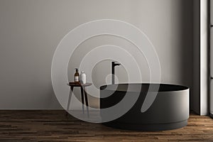 Modern oval black bathtub is standing in front gray wall and stool in empty bathroom. Minimalist concept.