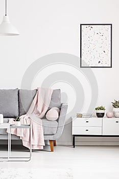 Modern poster on a white wall, grey sofa with blanket and cabinet in a living room interior. Real photo