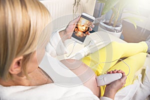 Modern pregnant woman sitting on a white couch at home relaxing and doing by herself digital 3D ultrasound
