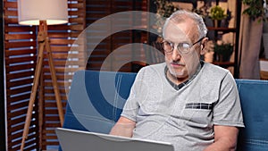 Modern retired man surfing internet by laptop at home in evening, portrait shot of retiree in room