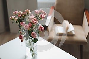 Modern room interior with armchair, book and fresh roses on table. Interior of beautiful living room decorated with flowers