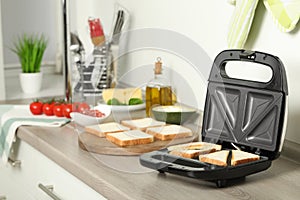 Modern sandwich maker with bread slices on wooden table in kitchen