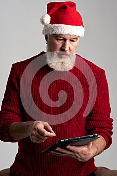 Modern Santa. Focused middle aged man with beard wearing Christmas hat and red sweater using digital tablet while