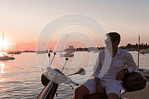 A modern senior businessman in casual clothes sitting on a scooter by the sea and enjoys the sunrise. Selective focus