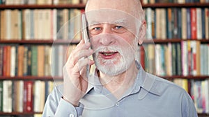 Modern senior man at home talking on cellphone in library. Bookcase bookshelves in background