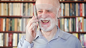 Modern senior man at home talking on cellphone in library. Bookcase bookshelves in background