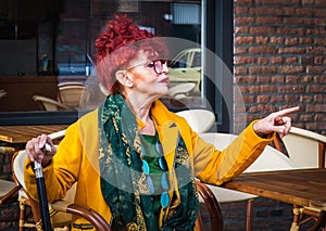 Fashionable woman in age sits in terrace of cafe and points with finger-modern lifestyle of pensionary, vacation or free time photo