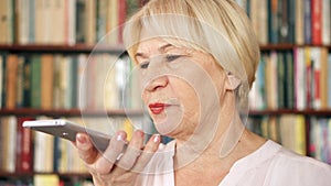 Modern senior woman at home talking on cellphone in library. Bookcase bookshelves in background