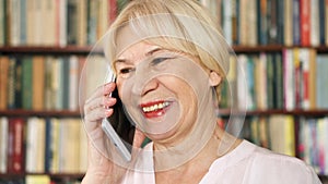 Modern senior woman at home talking on cellphone in library. Bookcase bookshelves in background