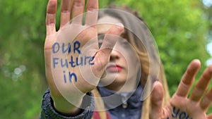 Modern serious girl with long dreadlocks is showing hands with written slogan `Our future in your hands` on green tree