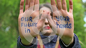 Modern serious girl with long dreadlocks is showing hands with written slogan `Our future in your hands` on green tree