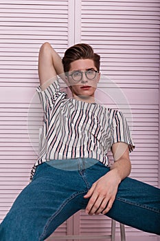 Modern sexy young man in fashionable youth wear in stylish glasses is resting on a chair near a wooden pink wall int the studio.