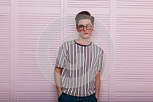 Modern sexy young man hipster in fashionable glasses with a hairstyle in a trendy striped t-shirt stand near a vintage wooden pink
