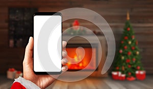 Modern smart phone mockup in Santa Claus hand with fireplace and Christmas tree with decorations and gift in background