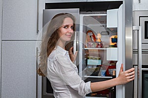 Modern smiling woman standing in the kitchen and open refrigerators door