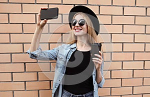 Modern smiling young woman taking selfie picture by smartphone with coffee cup wearing a black round hat, jeans jacket