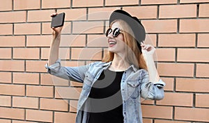 Modern smiling young woman taking selfie picture by smartphone wearing a black round hat, jeans jacket on brick wall