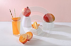 Modern still life with citrus fruits, orange juice and oranges, mangoes, pomegranates on a stand and podiums on a pink background