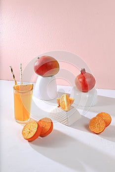 Modern still life with citrus, orange juice and oranges, pomegranate and mango on stand and podiums against pink background with