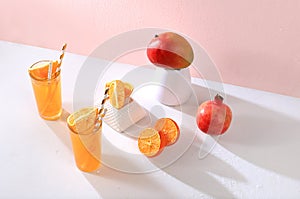Modern still life with citrus, orange juice and oranges, pomegranate and mango on stand and podiums on pink background with long