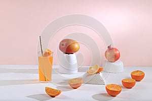 Modern still life with citrus, orange juice and oranges, pomegranate and mango on stand and podiums on pink background with long