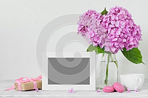 Modern still life with pink hydrangea flowers, photo frame, coffee cup and gift box on white wooden table
