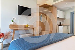Modern style interior of small studio apartment. Hotel room with white kitchen, blue bedroom, pine wooden wardrobe, flat TV