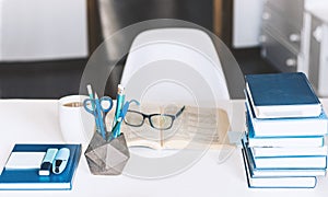 Modern stylish office work place with open book, glasses, office supplies and books, desk work concept in white and blue colors