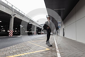 Modern stylish young hipster man in a black leather jacket in jeans in boots in sunglasses walks on the street near the parking