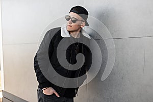 Modern stylish young hipster man in a stylish black jacket in a fashionable cap in trendy dark sunglasses in vintage gray jeans