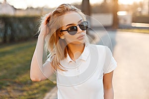 Modern stylish young hipster woman in trendy white polo t-shirt in black sunglasses stand and posing