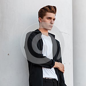 Modern stylish young man in trendy black shirt in a t-shirt with a fashionable hairstyle poses near a white vintage building