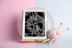 Modern tablet wrapped in thread, yarn ball and knitting needles on color background. Internet addiction concept