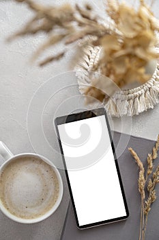 Modern tabletop with with smartphone mockup, cup of coffee, dried plants and notebook on textured white background. Flat lay,