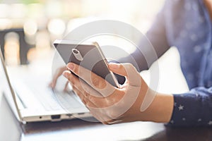 Modern technology and lifestyle concept. A close-up of woman`s hands holding smartphone and typing something at her laptop. A woma