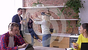 Modern technology in office, young female with Virtual reality helmet plays games while collaborators eat and chat while