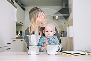 Modern tired mother and little child after sleepless night. Exhausted woman with baby is sitting with coffee in kitchen. Life of